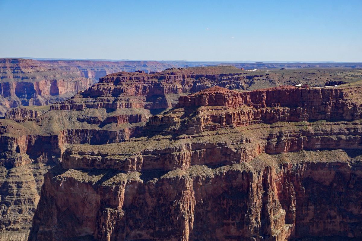 Wide view of layered red and tan canyon cliffs and flat-topped mesas stretching toward a hazy horizon under a clear blue sky.