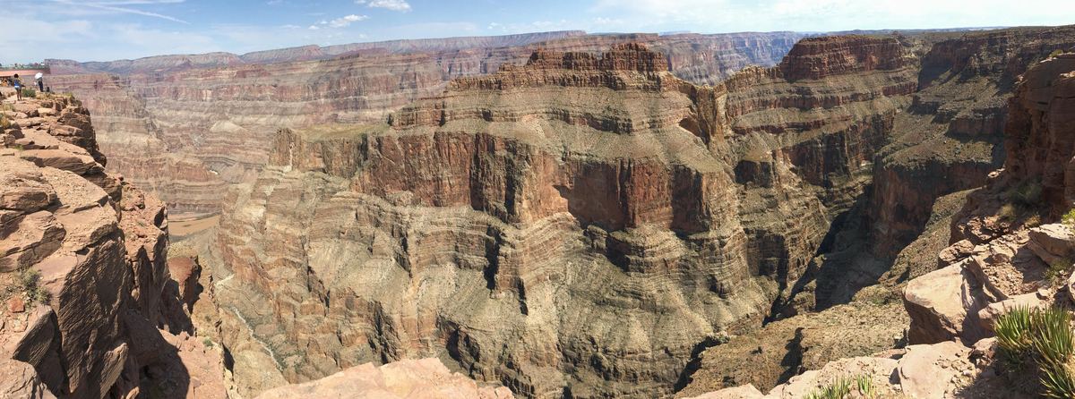 Wide panoramic view of a deep canyon with layered red and tan rock cliffs under a mostly clear blue sky.
