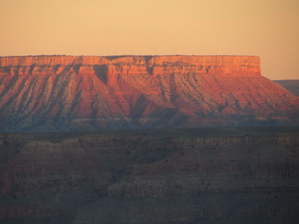 A flat-topped red rock mesa lit in warm orange light above darker, shadowed canyon slopes under a soft peach sky.