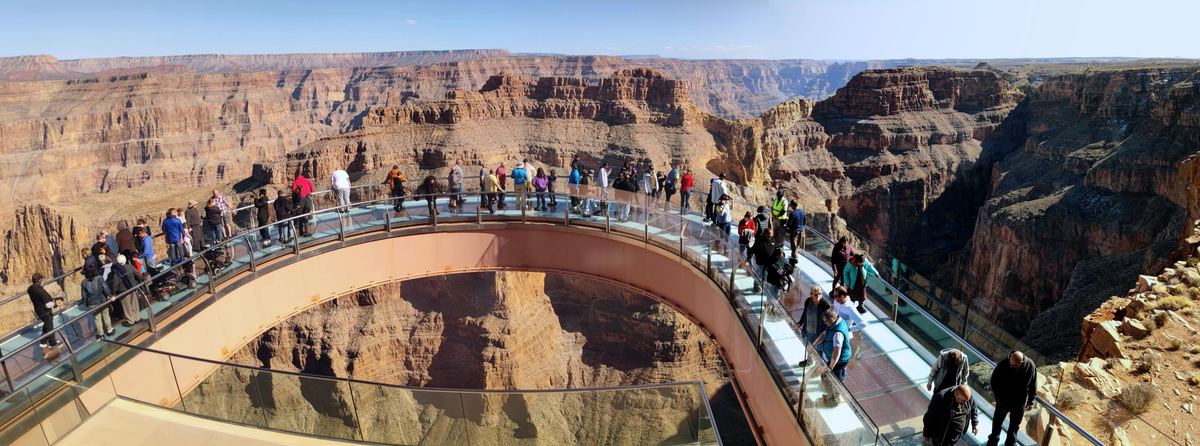 Visitors stand on a curved glass-floored walkway jutting out over a deep canyon with layered red and tan rock walls under a clear sky.