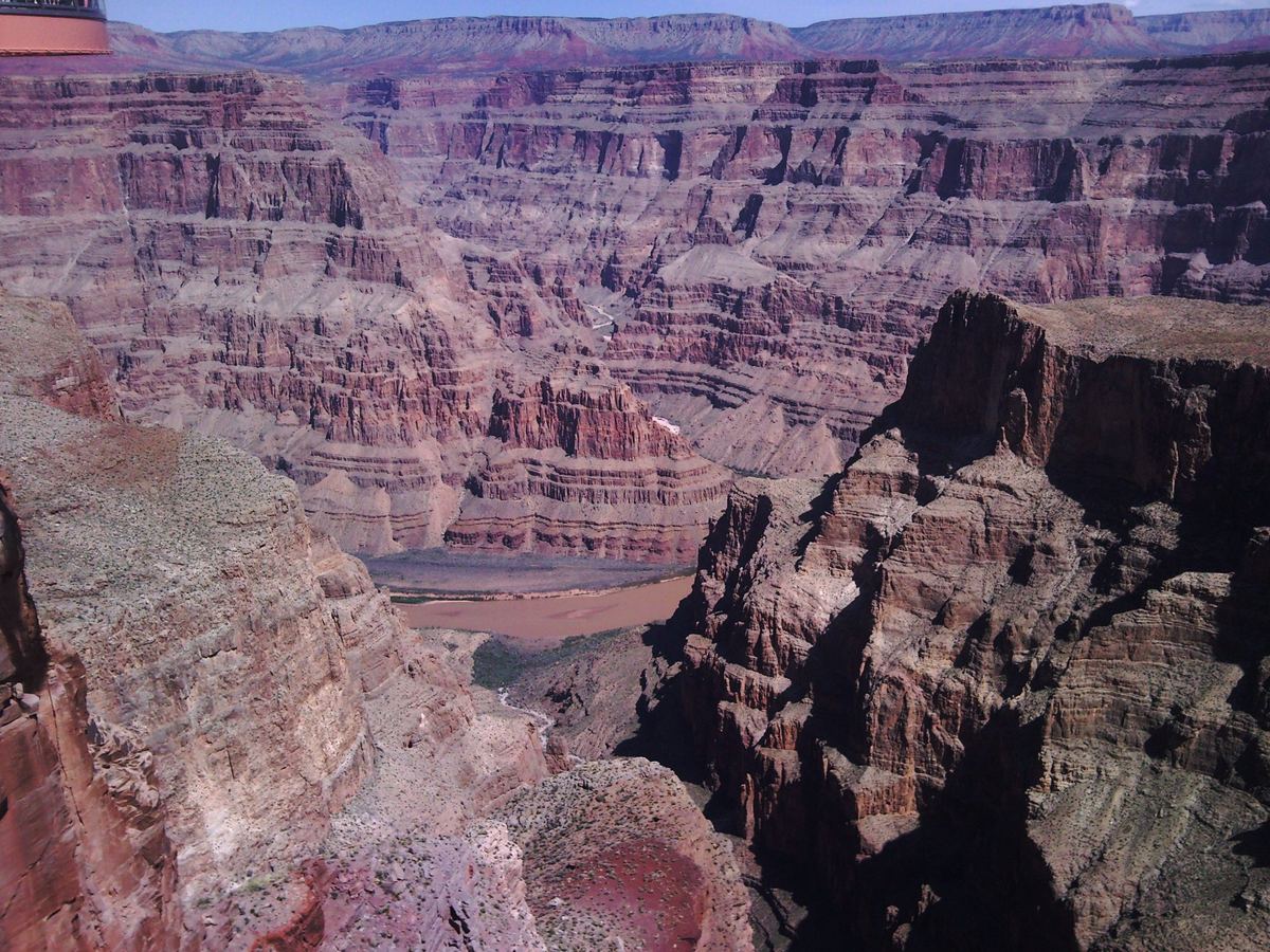 Wide view of a deep canyon with layered red and tan rock cliffs and a muddy river at the bottom.