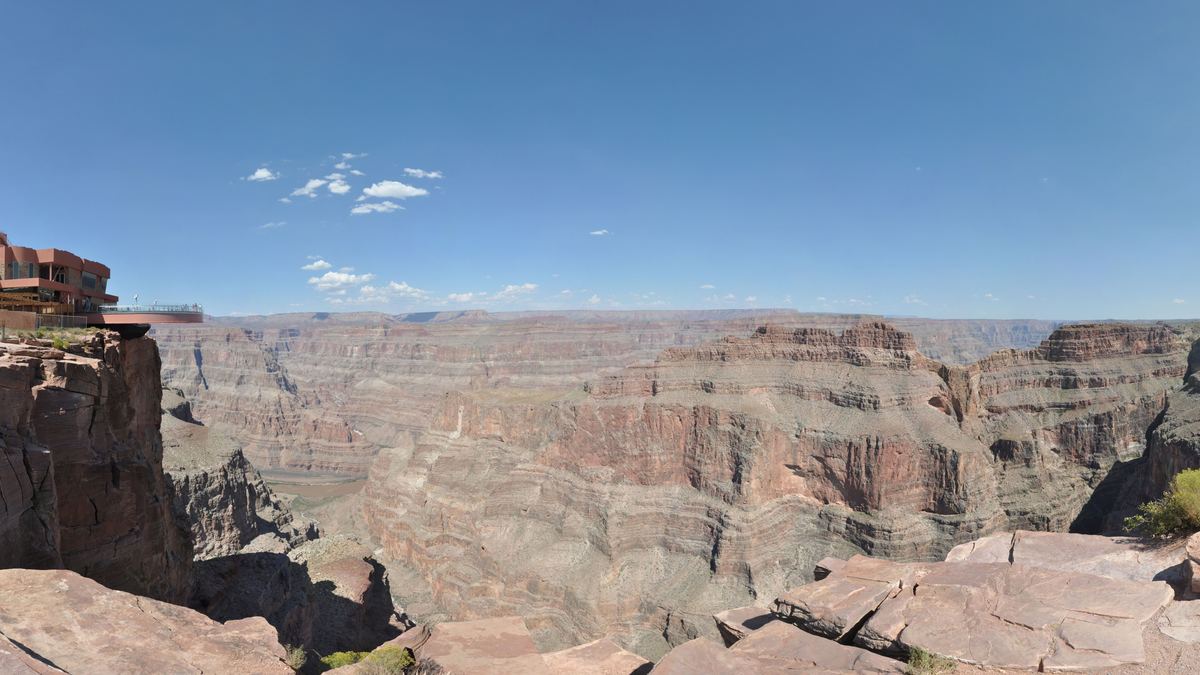 Wide panorama of a deep canyon with layered red and tan rock walls under a blue sky, with a building on the rim at left and rocky ledges in the foreground.