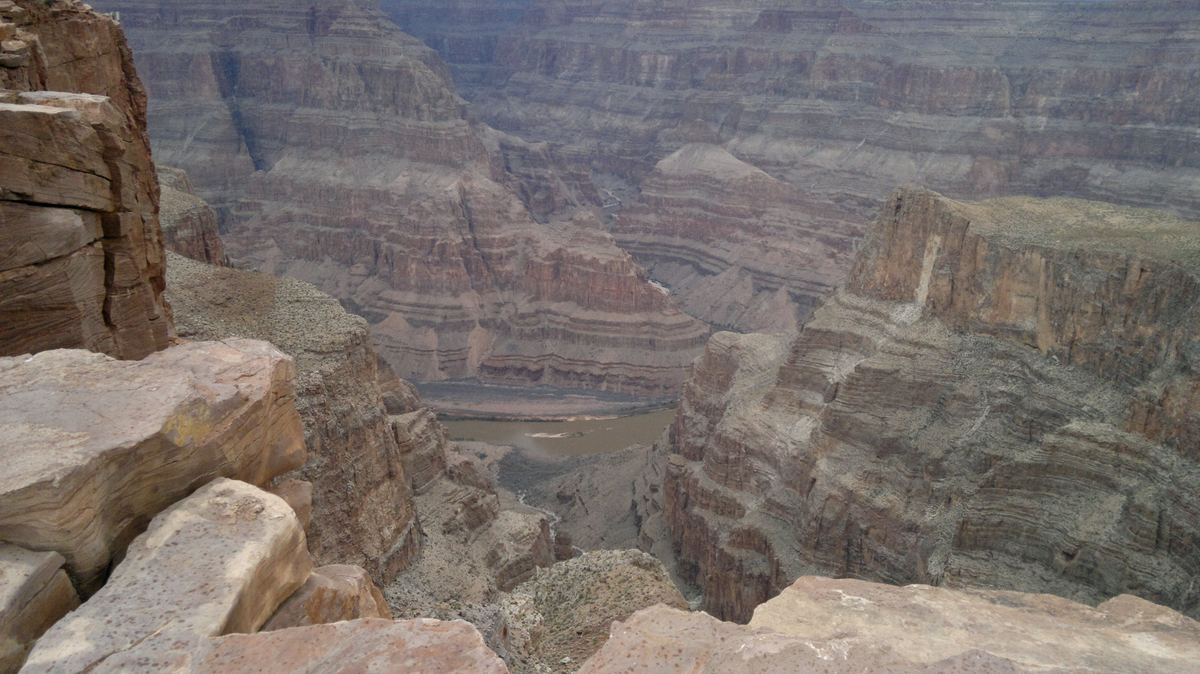 View from a rocky ledge into a deep canyon with layered cliffs and a river winding along the canyon floor under a hazy sky.