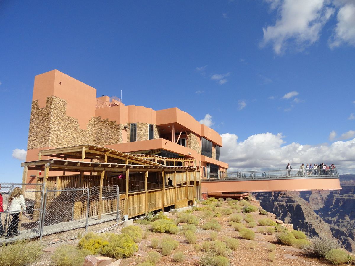 A terracotta-colored visitor building with stone walls and a wooden deck, beside a curved walkway extending over a canyon under a blue sky with clouds.