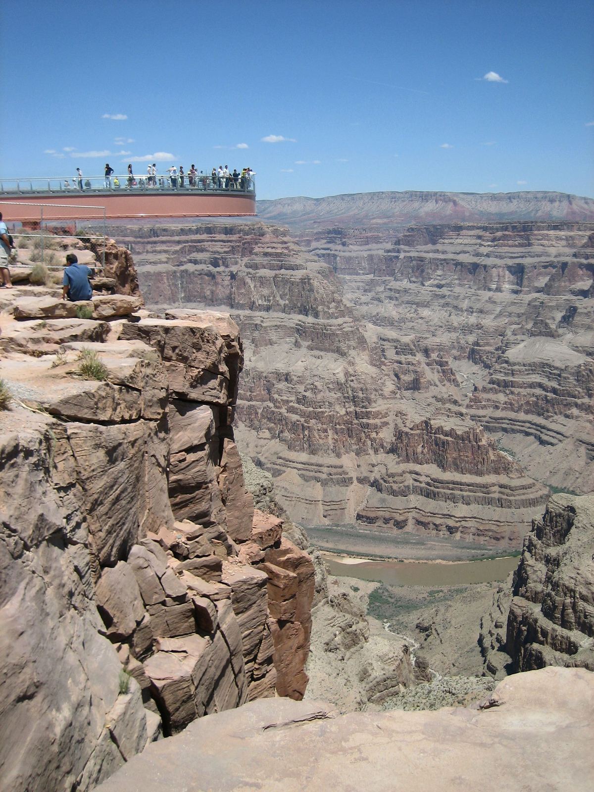 A rocky cliff edge overlooks a deep layered canyon, with a curved walkway platform and a crowd of small visitors on the far rim under a blue sky.